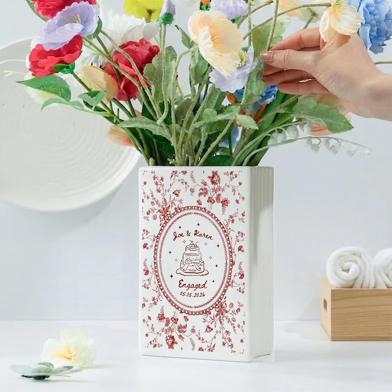 May include: A rectangular white ceramic vase featuring a red floral pattern and a central oval frame. The frame contains a line drawing of a tiered cake with the text Joe & Karen, Engaged, and the date 05.15.2026 printed in red ink.