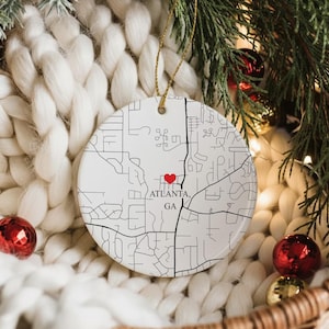 Puede incluir: Adorno navide&ntilde;o de cer&aacute;mica blanca con un mapa de Atlanta, GA, marcado con un coraz&oacute;n rojo. El adorno cuelga de un hilo dorado y est&aacute; rodeado de decoraciones festivas, incluyendo adornos rojos y dorados y ramas de hoja perenne.