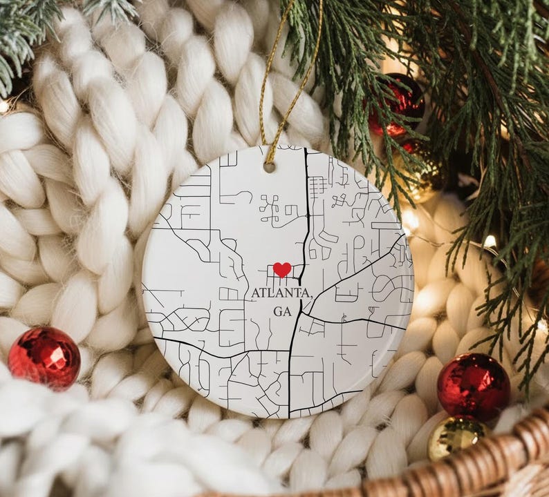 Puede incluir: Adorno navide&ntilde;o de cer&aacute;mica blanca con un mapa de Atlanta, GA, marcado con un coraz&oacute;n rojo. El adorno cuelga de un hilo dorado y est&aacute; rodeado de decoraciones festivas, incluyendo adornos rojos y dorados y ramas de hoja perenne.