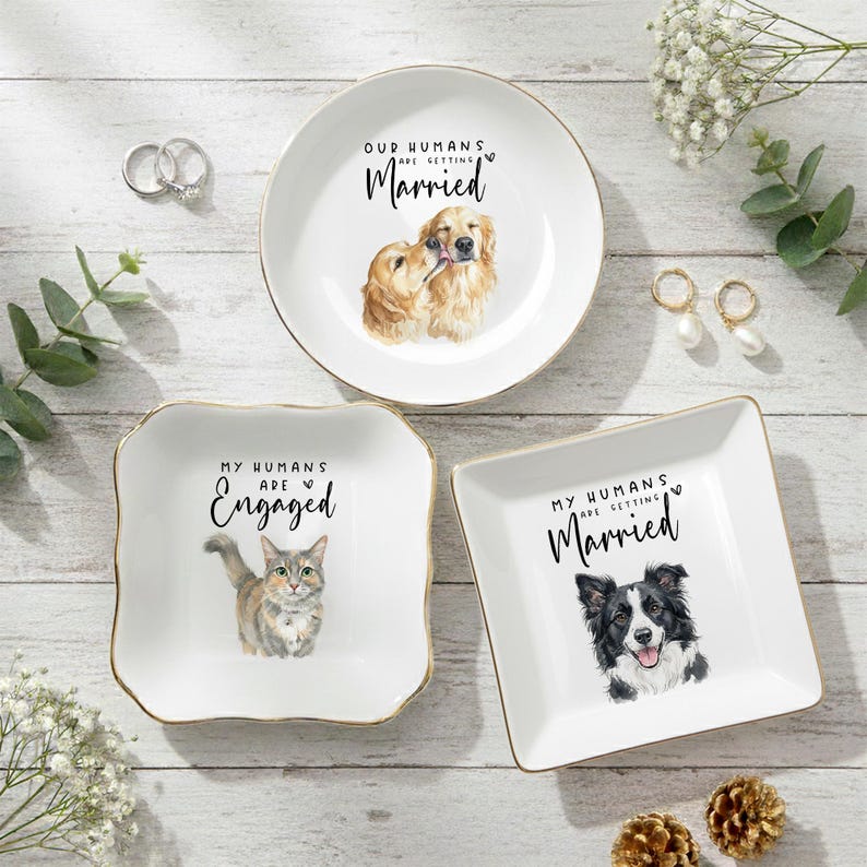 May include: Three white ceramic ring dishes with gold rims. One dish features a Golden Retriever illustration and the text "Our Humans Are Getting Married." Another has a cat illustration and the text "My Humans Are Engaged." The third dish has a Border Collie illustration and the text "My Humans Are Getting Married."