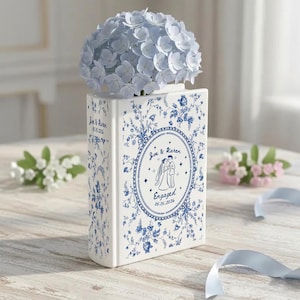 May include: A white ceramic book-shaped vase with blue floral patterns. The front features an oval frame with an illustration of a couple and the text "Joe & Karen Engaged 05.15.2026". The spine displays "Joe & Karen 05.15.2026". A bouquet of light blue hydrangea flowers sits inside.
