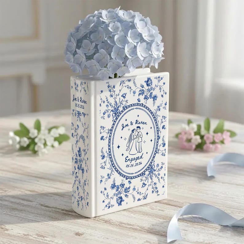 May include: A white ceramic book-shaped vase with blue floral patterns. The front features an oval frame with an illustration of a couple and the text "Joe & Karen Engaged 05.15.2026". The spine displays "Joe & Karen 05.15.2026". A bouquet of light blue hydrangea flowers sits inside.