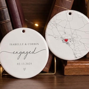Puede incluir: Dos adornos redondos de cer&aacute;mica blanca. Uno presenta un mapa con un coraz&oacute;n rojo, el otro dice "Isabelle & Corbin engaged 02.13.2025" en escritura negra. Ambos tienen un agujero para colgar. Los adornos se exhiben cerca de libros.