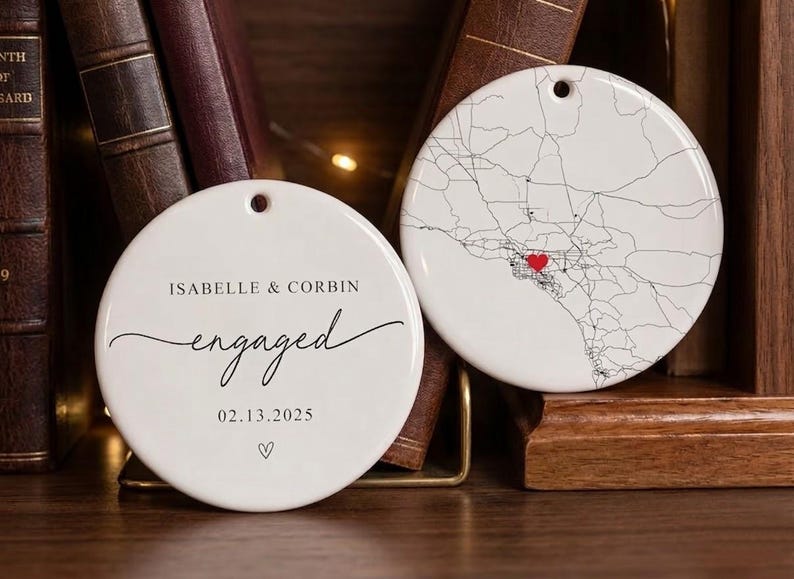 Puede incluir: Dos adornos redondos de cer&aacute;mica blanca. Uno presenta un mapa con un coraz&oacute;n rojo, el otro dice "Isabelle & Corbin engaged 02.13.2025" en escritura negra. Ambos tienen un agujero para colgar. Los adornos se exhiben cerca de libros.