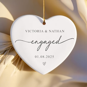 Puede incluir: Adorno blanco en forma de coraz&oacute;n con una cuerda dorada para colgar. Tiene los nombres "VICTORIA & NATHAN", la palabra "engaged" en cursiva y la fecha "01.08.2025". Un peque&ntilde;o coraz&oacute;n en la parte inferior.