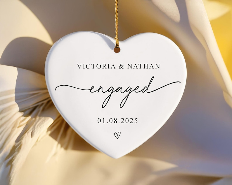 Puede incluir: Adorno blanco en forma de coraz&oacute;n con una cuerda dorada para colgar. Tiene los nombres "VICTORIA & NATHAN", la palabra "engaged" en cursiva y la fecha "01.08.2025". Un peque&ntilde;o coraz&oacute;n en la parte inferior.