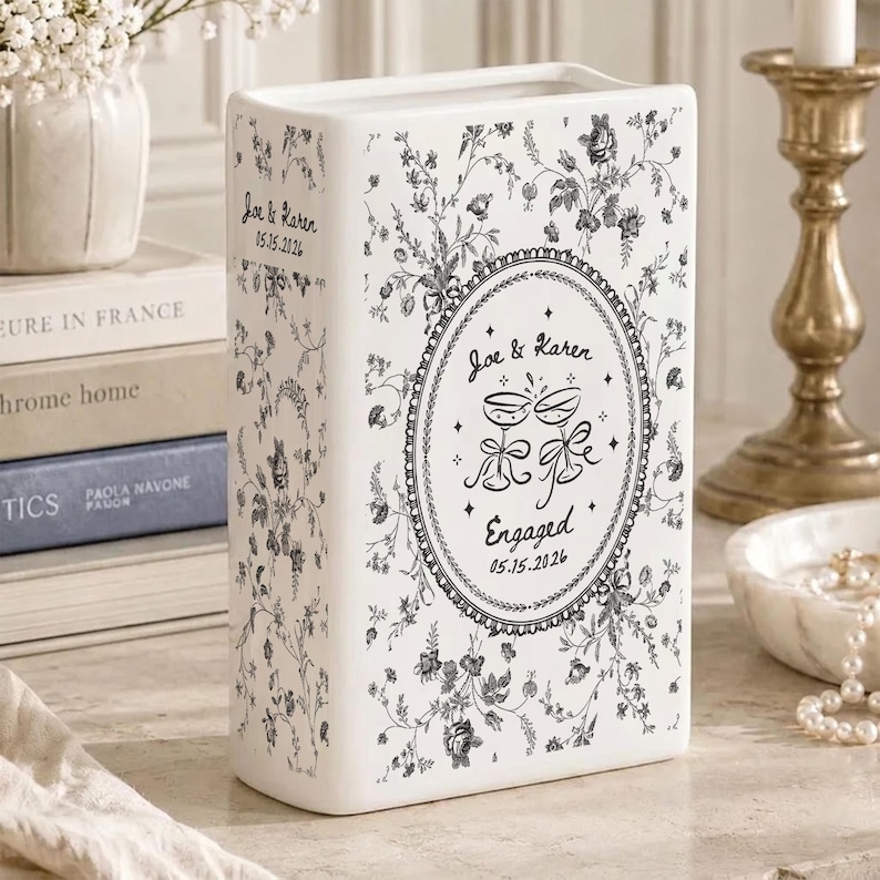 May include: A white ceramic book-shaped vase featuring a black floral toile pattern. The front displays an oval frame with the text "Joe & Karen", "Engaged", and "05.15.2026" alongside a graphic of two clinking champagne glasses with ribbons.