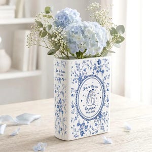 May include: A rectangular white ceramic vase with blue floral patterns, featuring a central oval frame with an illustration of a couple and the text Joe & Karen Engaged 05.15.2026. The side displays the text Joe & Karen 05.15.2026. Filled with blue hydrangeas.