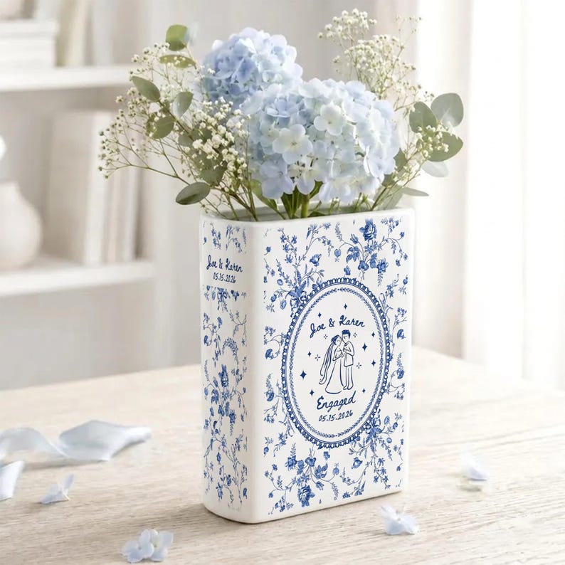 May include: A rectangular white ceramic vase with blue floral patterns, featuring a central oval frame with an illustration of a couple and the text Joe & Karen Engaged 05.15.2026. The side displays the text Joe & Karen 05.15.2026. Filled with blue hydrangeas.