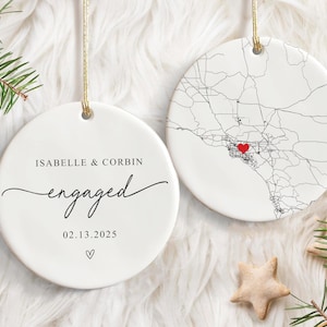 May include: Two white ceramic ornaments with gold string hangers. One ornament reads "ISABELLE & CORBIN engaged 02.13.2025" with a heart symbol. The other features a map design with a red heart.