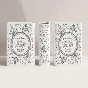 May include: Two rectangular white ceramic vases and one narrow central vase, all featuring a black floral pattern. The front of the vases displays an oval frame with two clinking glasses, the text Joe & Karen, Engaged, and 05.15.2026.
