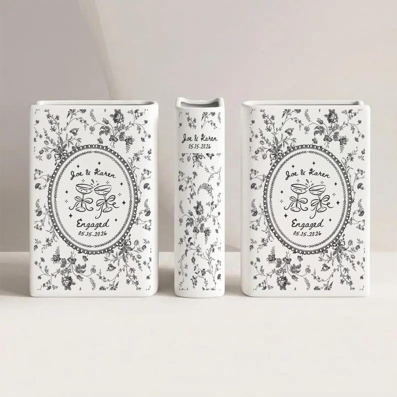 May include: Two rectangular white ceramic vases and one narrow central vase, all featuring a black floral pattern. The front of the vases displays an oval frame with two clinking glasses, the text Joe & Karen, Engaged, and 05.15.2026.