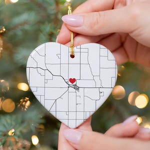Puede incluir: Adorno de cer&aacute;mica en forma de coraz&oacute;n con un dise&ntilde;o de mapa y un coraz&oacute;n rojo que marca una ubicaci&oacute;n. El adorno blanco tiene l&iacute;neas negras que representan las calles y un coraz&oacute;n rojo. Se adjunta una cuerda dorada para colgar.