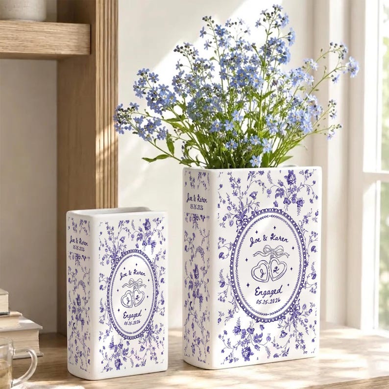 May include: Two rectangular white ceramic vases featuring a blue floral pattern. The vases are personalized with the text 'Joe & Karen', 'Engaged', and the date '05.15.2026' inside an oval frame with two heart-shaped rings and a ribbon bow.