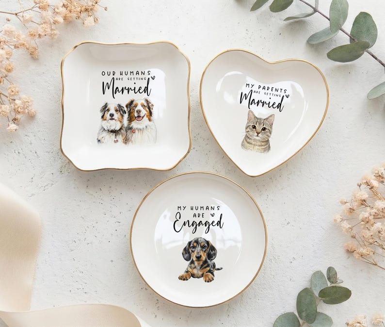 May include: Three white ceramic ring dishes with gold rims. One square dish features two dogs and the text "Our Humans Are Getting Married." A heart-shaped dish has a cat and the text "My Parents Are Getting Married." A round dish shows a dachshund and the text "My Humans Are Engaged."
