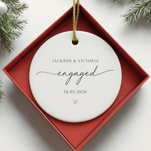 Puede incluir: Adorno de compromiso de cer&aacute;mica blanca con los nombres "Jackson & Victoria" y la palabra "engaged" en escritura cursiva. La fecha "26.02.2026" y un coraz&oacute;n est&aacute;n debajo. El adorno est&aacute; en una caja roja con una cuerda dorada.