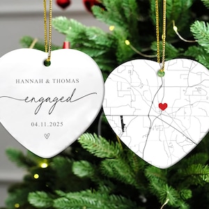 Puede incluir: Dos adornos blancos de cer&aacute;mica en forma de coraz&oacute;n, colgados de cordones dorados. Uno dice "Hannah & Thomas engaged 04.11.2025" en escritura negra. El otro presenta un mapa en blanco y negro con un coraz&oacute;n rojo.
