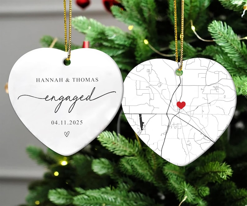 Puede incluir: Dos adornos blancos de cer&aacute;mica en forma de coraz&oacute;n, colgados de cordones dorados. Uno dice "Hannah & Thomas engaged 04.11.2025" en escritura negra. El otro presenta un mapa en blanco y negro con un coraz&oacute;n rojo.