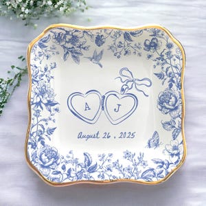 May include: Square ceramic dish with a gold rim and blue floral design. The center features two heart-shaped lockets with the letters "A" and "J", and the date "August 26, 2025". Decorative butterflies and hummingbirds are also present.
