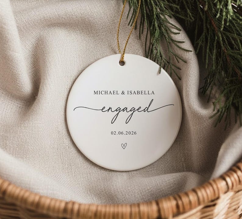 Puede incluir: Adorno de compromiso de cer&aacute;mica blanca con los nombres "MICHAEL & ISABELLA" y la palabra "engaged" en escritura cursiva. La fecha "02.06.2026" y un s&iacute;mbolo de coraz&oacute;n tambi&eacute;n est&aacute;n presentes. El adorno cuelga de un hilo dorado.