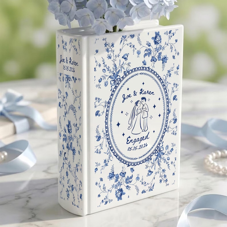 May include: A white ceramic book-shaped vase with blue floral patterns. The front features an oval frame with a line drawing of a couple, the text "Joe & Karen" and "Engaged 05.15.2026". The spine also displays "Joe & Karen 05.15.2026".