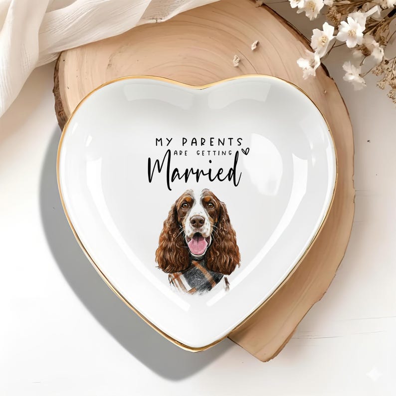 May include: Heart-shaped white ceramic trinket dish with a gold rim. The dish features a dog illustration and the text "MY PARENTS ARE GETTING Married". The dog has brown and white fur and is wearing a plaid scarf.