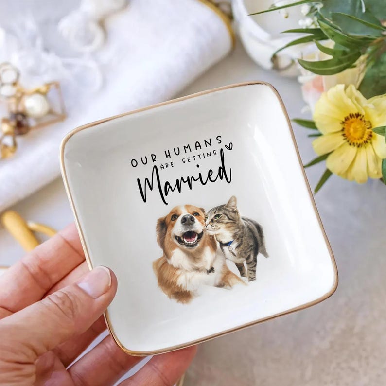 May include: Square white ceramic trinket dish with gold trim. Features a watercolor illustration of a dog and cat with the text "OUR HUMANS ARE GETTING Married". A charming gift for pet lovers.