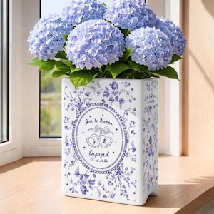 May include: A rectangular white ceramic vase featuring a blue floral pattern and a central oval emblem with the text Joe & Karen, Engaged, 05.15.2026, and two heart icons. The vase is filled with a large bouquet of vibrant light blue hydrangea flowers.