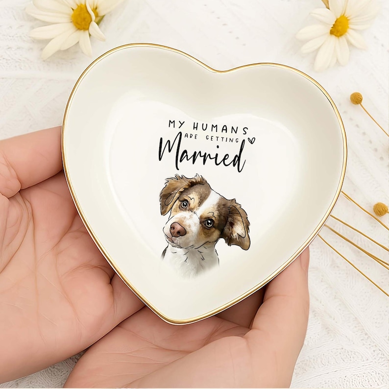 May include: A heart-shaped, off-white ceramic trinket dish with a gold rim. The dish features a watercolor illustration of a dog and the text "MY HUMANS are getting Married". A charming gift for a pet owner.