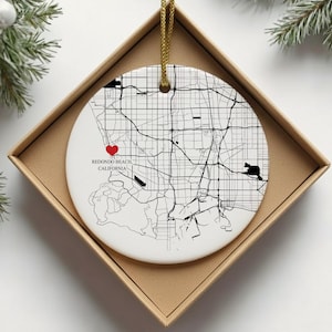 Puede incluir: Adorno navide&ntilde;o de cer&aacute;mica blanca con un mapa en blanco y negro de Redondo Beach, California. Un coraz&oacute;n rojo marca una ubicaci&oacute;n en el mapa. El adorno est&aacute; en una caja de cart&oacute;n marr&oacute;n con un cord&oacute;n dorado.