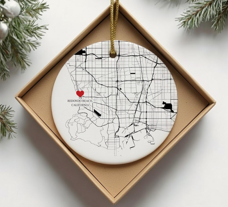 Puede incluir: Adorno navide&ntilde;o de cer&aacute;mica blanca con un mapa en blanco y negro de Redondo Beach, California. Un coraz&oacute;n rojo marca una ubicaci&oacute;n en el mapa. El adorno est&aacute; en una caja de cart&oacute;n marr&oacute;n con un cord&oacute;n dorado.
