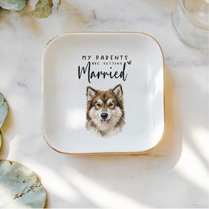 May include: A white square ceramic trinket dish with gold trim. The dish features the text "MY PARENTS ARE GETTING Married" and a watercolor illustration of a dog. The dog has brown and white fur and a happy expression.