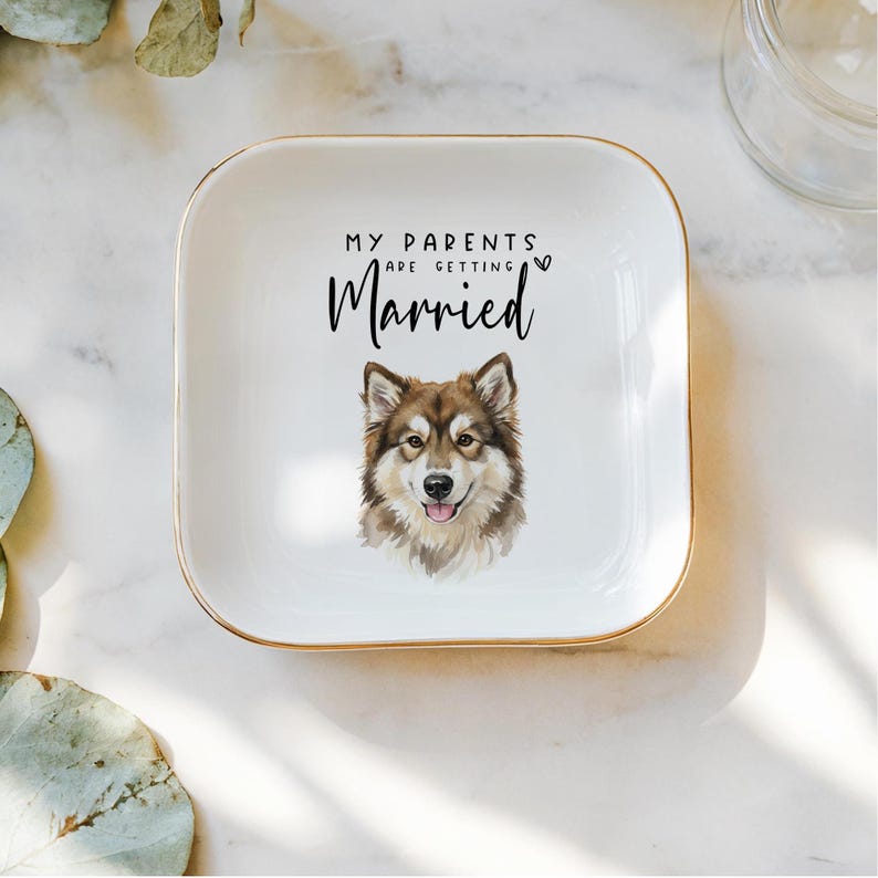 May include: A white square ceramic trinket dish with gold trim. The dish features the text "MY PARENTS ARE GETTING Married" and a watercolor illustration of a dog. The dog has brown and white fur and a happy expression.