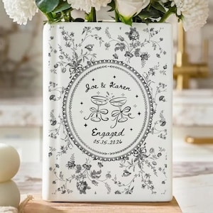 May include: A rectangular white ceramic vase featuring a black floral toile pattern. The center displays an oval frame with the text Joe & Karen, a graphic of two clinking champagne glasses, the word Engaged, and the date 05.15.2026.