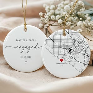 May include: Two white ceramic ornaments with gold string hangers. One ornament says "SAMUEL & ELORA engaged 01.05.2026" with a heart. The other features a map of Newport Beach, CA, with a red heart.