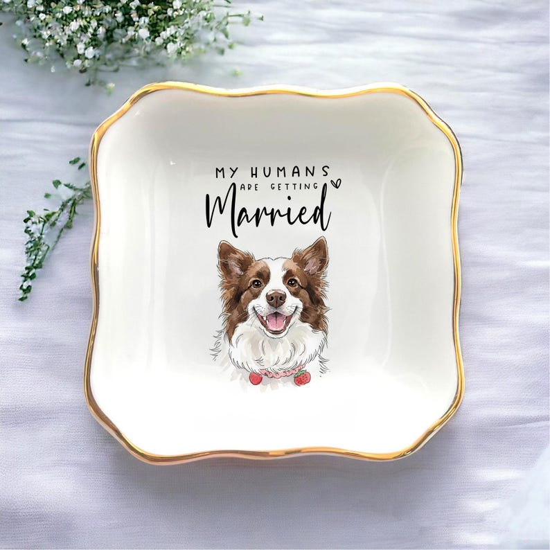 May include: A white ceramic trinket dish with a gold rim and a scalloped edge. The dish features a smiling dog illustration and the text "MY HUMANS ARE GETTING Married". A charming wedding gift.