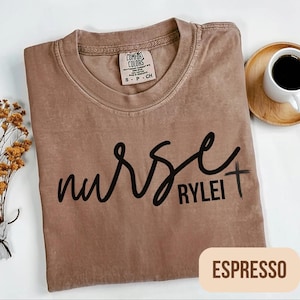 Puede incluir: Camiseta color espresso con la palabra "nurse" en cursiva y el nombre "Rylei" debajo, con una peque&ntilde;a cruz. La camiseta es de la marca Comfort Colors, talla small.