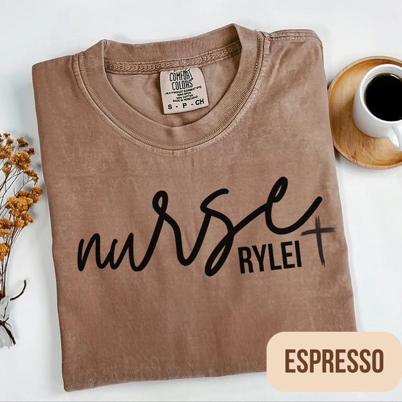 Puede incluir: Camiseta color espresso con la palabra "nurse" en cursiva y el nombre "Rylei" debajo, con una peque&ntilde;a cruz. La camiseta es de la marca Comfort Colors, talla small.