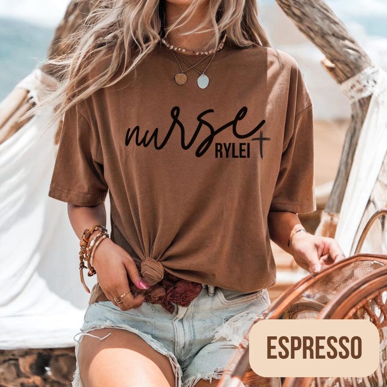 Puede incluir: Camiseta color espresso con la palabra "nurse" en cursiva y el nombre "Rylei" debajo. La camiseta est&aacute; anudada a la cintura. Es una camiseta de manga corta y cuello redondo.