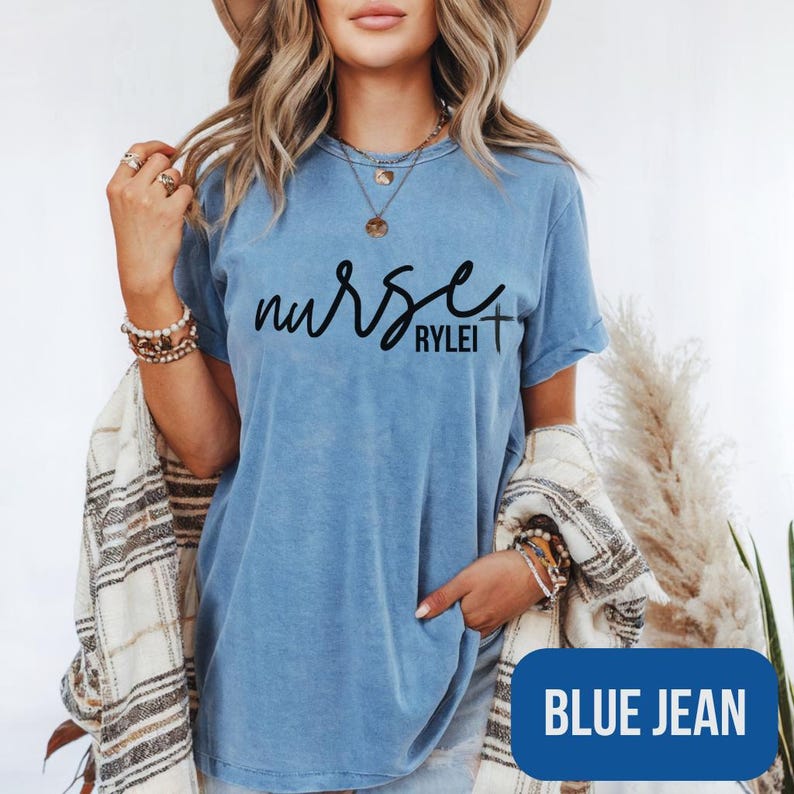 Puede incluir: Camiseta azul vaquero con la palabra "nurse" en cursiva, con el nombre "Rylei" debajo. La camiseta tiene un s&iacute;mbolo de cruz al final de la palabra "nurse". La camiseta es de estilo cuello redondo.