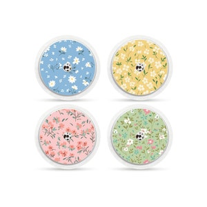 Könnte beinhalten: Vier runde, selbstklebende Aufkleber mit verschiedenen Blumenmustern. Die Aufkleber sind blau, gelb, rosa und grün mit weißen Blumen und grünen Blättern.