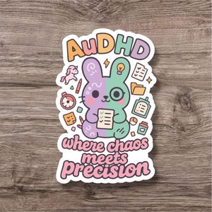 Könnte beinhalten: Aufkleber mit einem Cartoon-Hasen-Design, aufgeteilt in lila und grüne Hälften, der eine Checkliste hält. Der Text lautet "AuDHD where chaos meets precision." Das Design umfasst kleine Illustrationen eines Einhorns, einer Glühbirne und anderer Symbole.