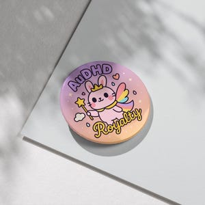Peut inclure: Un bouton rond aux couleurs pastel avec un lapin de dessin animé portant une couronne et tenant une baguette étoilée. Le bouton porte le texte "AUDHD Royalty" dans une police ludique, avec un fond dégradé rose, orange et violet.