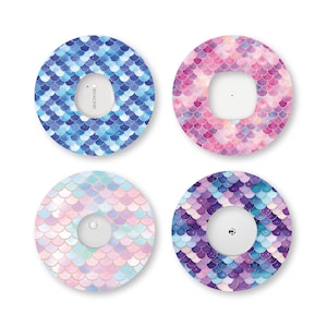 May include: Four circular adhesive patches for medical sensors, each featuring a distinct mermaid scale pattern. The top left patch is blue, the top right is pink, the bottom left is pastel, and the bottom right is purple and blue.
