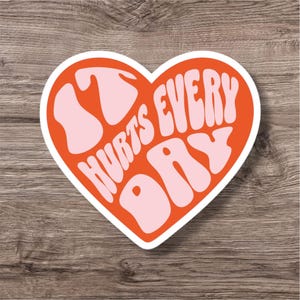 Op de afbeelding: Een hartvormige sticker met de tekst "It Hurts Every Day" in een retro lettertype. Het hart is oranje met een witte rand en de tekst is lichtroze. De sticker ligt op een houten ondergrond.