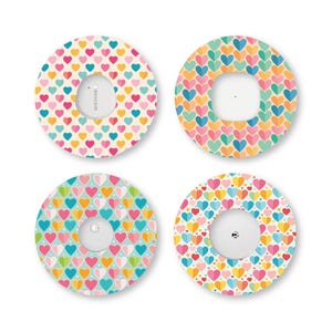 May include: Four circular adhesive patches for medical sensors, each featuring a colourful heart pattern. The designs include various combinations of pink, yellow, blue, and orange hearts on cream, teal, and white backgrounds, with central cutouts.