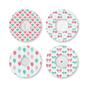 Puede incluir: Cuatro parches adhesivos circulares para dispositivos médicos, cada uno con un patrón distinto: corazones rosas y verde azulado, cerezas rojas sobre fondo azul claro, globos rosas y verde azulado, y lazos rosas y verde azulado sobre azul.
