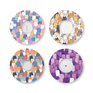 May include: Four circular adhesive patches for medical devices, each featuring a repeating pattern of cartoon cats. The designs include multicolored cats, brown and grey cats, blue and orange cats, and various shades of purple cats with white cats.