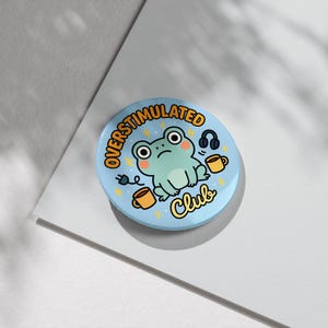 Peut inclure: Un bouton bleu clair avec les mots "OVERSTIMULATED Club" en jaune. Le bouton représente une grenouille de dessin animé avec un casque, deux tasses à café et un éclair.