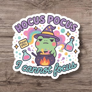 Op de afbeelding: Een sticker met een groene kikker met een heksenhoed, die in een ketel met vlammen roert. De tekst "Hocus Pocus I cannot focus" is in paars en teal. Het ontwerp bevat sterren, drankflessen en een regenboog.